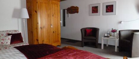 A bedroom with a large wooden wardrobe, a bed with a red blanket, a chair, a small table with a vase, and two framed pictures on the wall.