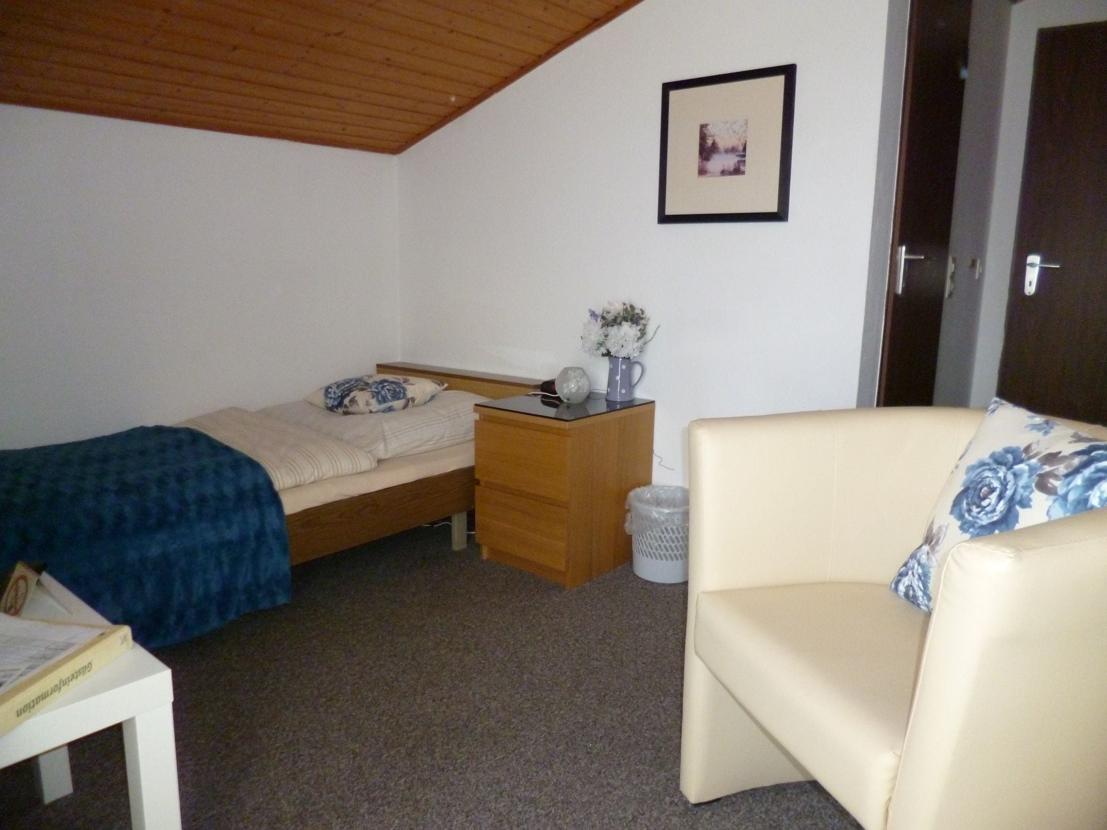 A small, sparsely furnished room with a bed, a dresser, a chair, and a picture on the wall.