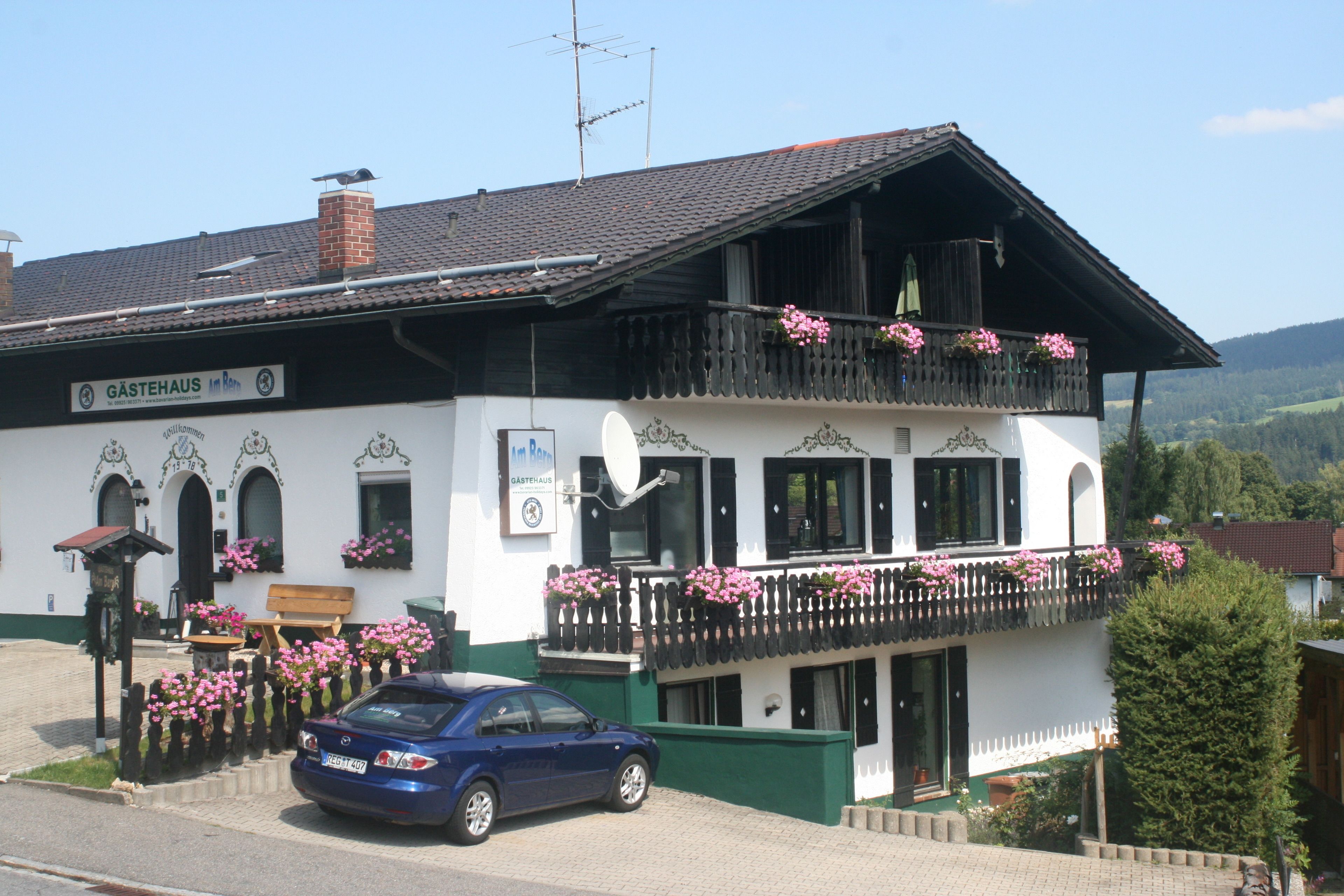 A white building with a balcony adorned with pink flowers, a blue car parked in front, and a sign reading "GÄSTEHAUS".