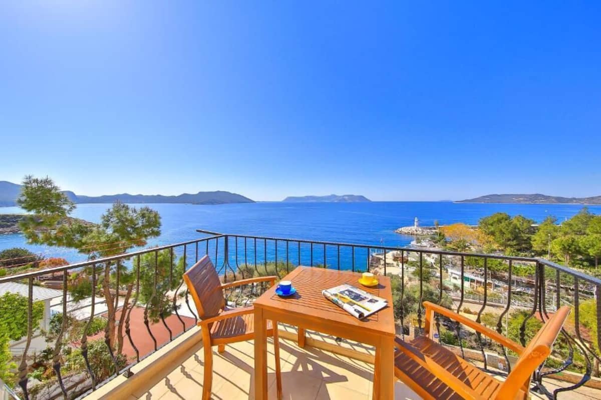 Un balcon avec une table et des chaises en bois, offrant une vue sur la côte, avec une eau d’un bleu limpide et des montagnes à l’horizon.