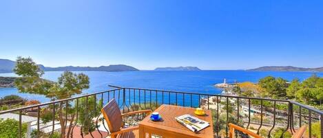A balcony with a wooden table and chairs, overlooking a coastal view with clear blue waters and distant mountains.