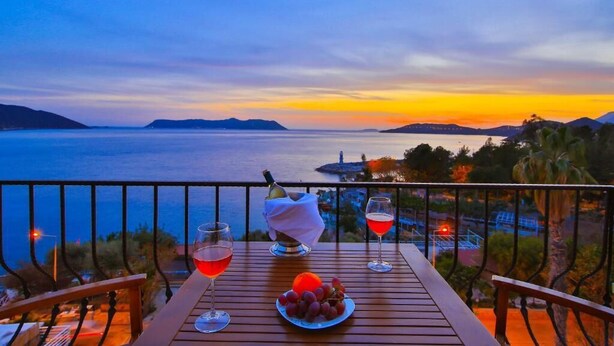 A wooden table with two glasses of red wine and a bowl of grapes, overlooking a scenic view of the sea and distant islands during sunset.