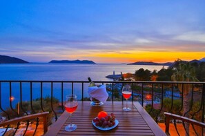A wooden table with two glasses of red wine and a bowl of grapes, overlooking a scenic view of the sea and distant islands during sunset. - Hotel Kayahan (Kas)