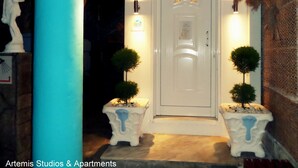 Una puerta blanca con un pequeño arco encima, flanqueada por dos macetas. A la izquierda hay una estatua y a la derecha un aplique de pared.