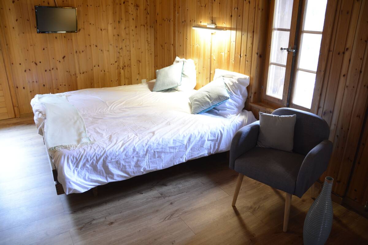 A room with a wooden paneled wall, a bed with white bedding, a grey armchair, and a large window with curtains.