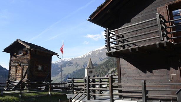 A wooden building with a staircase leading to a balcony, a flag, and a mountain in the background. - Studios du Valsorey (Bourg-Saint-Pierre)