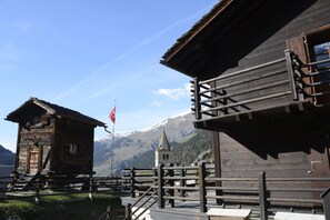 A wooden building with a staircase leading to a balcony, a flag, and a mountain in the background. - Studios du Valsorey (Bourg-Saint-Pierre)