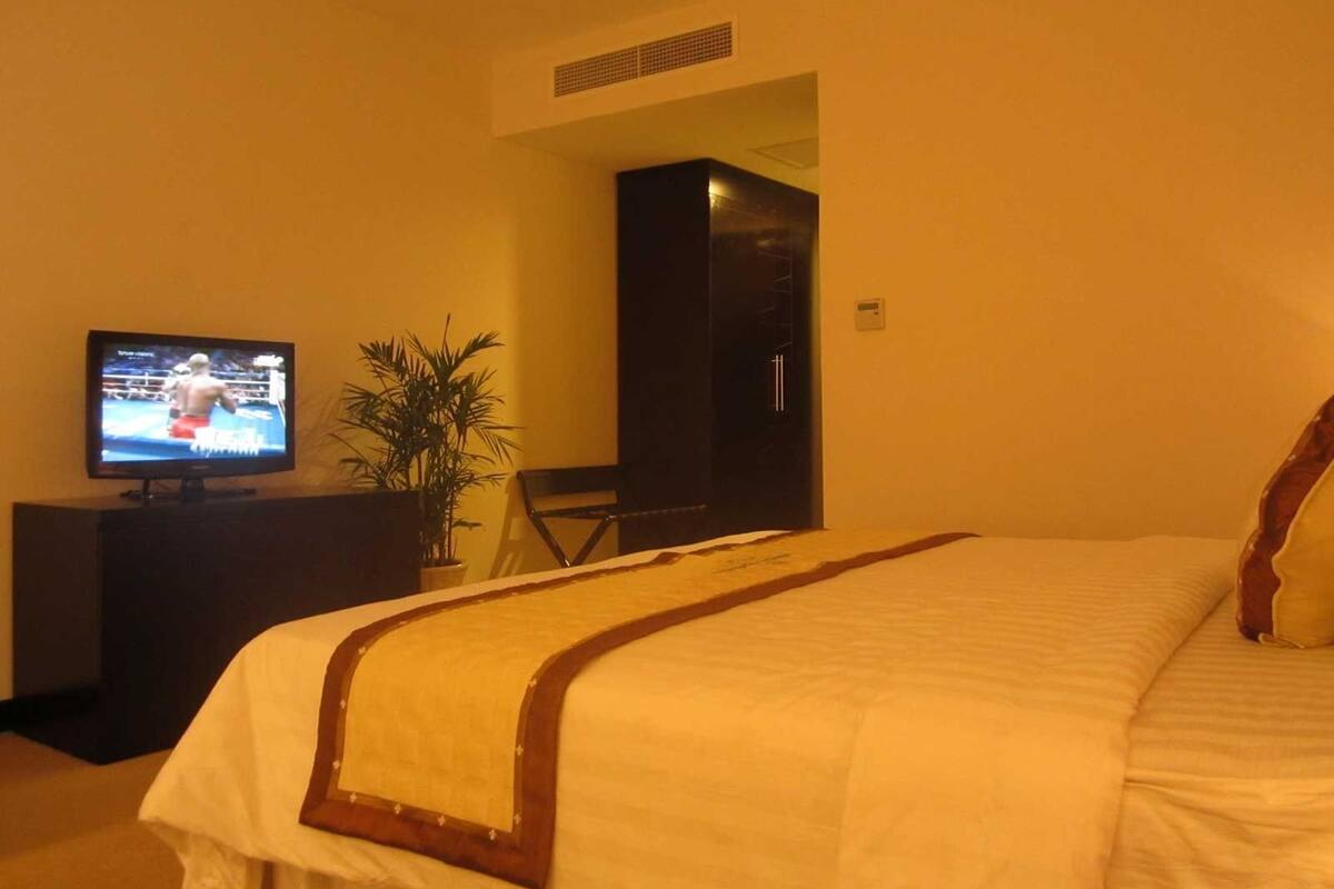 Une chambre d’hôtel avec un lit, un téléviseur diffusant un match de boxe, une plante en pot et une porte donnant sur une autre pièce.