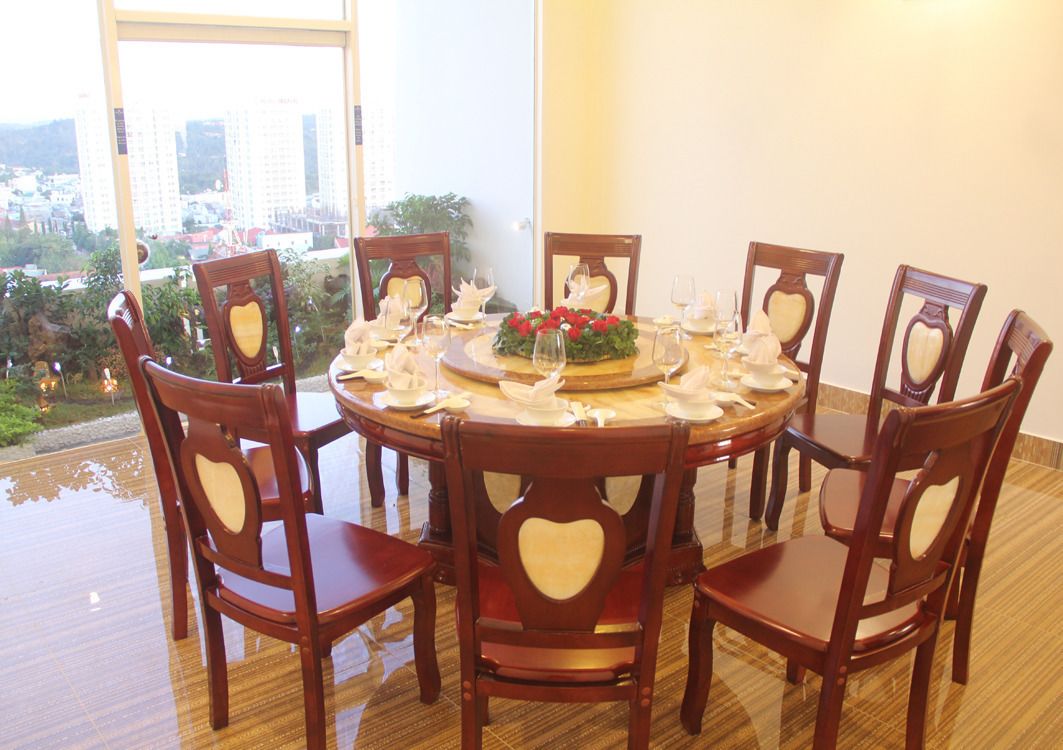 Una mesa de comedor redonda con ocho sillas de madera, cada una con respaldo en forma de corazón. La mesa está preparada para una comida con platos, cubiertos y un centro de mesa con flores rojas.