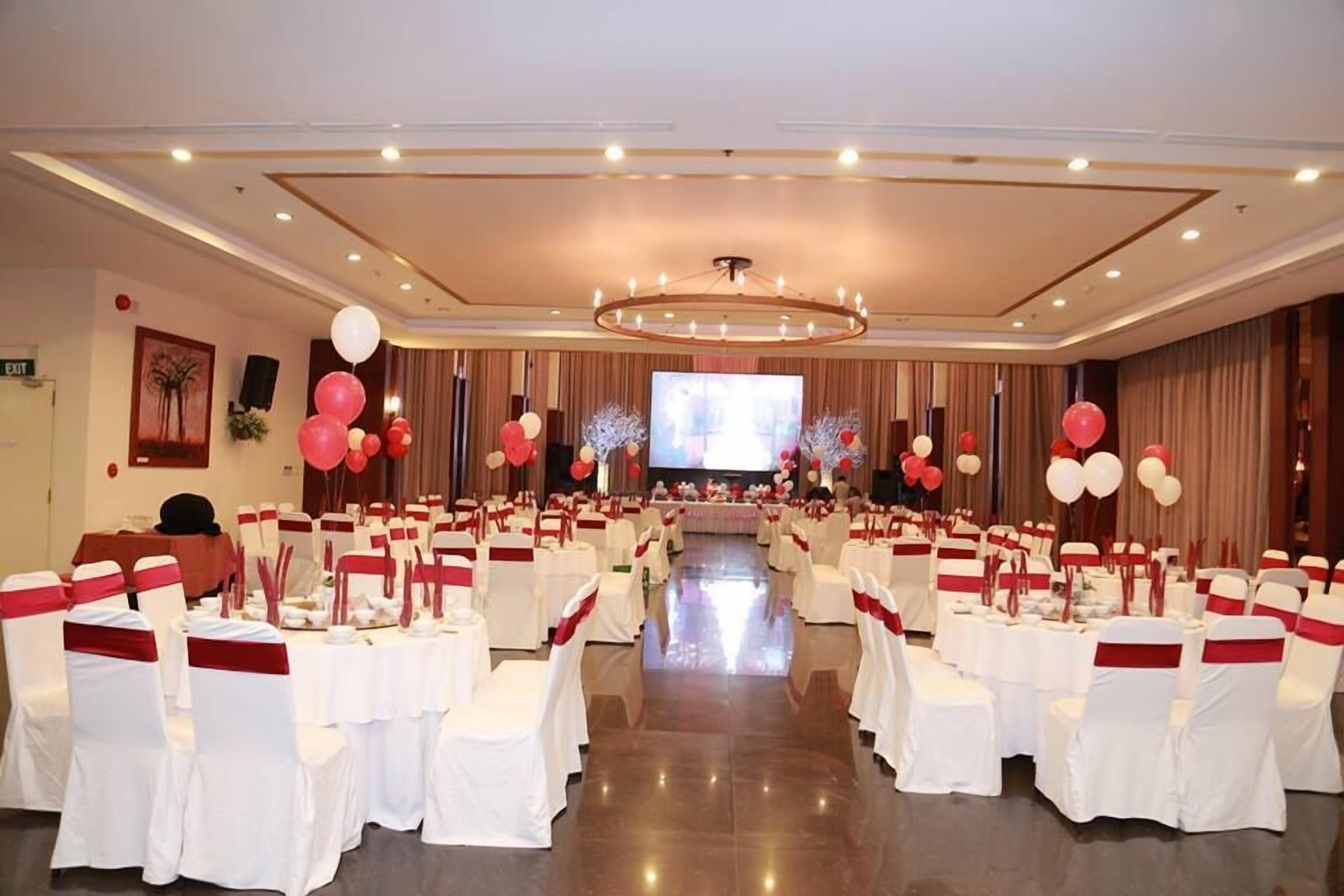 Un salón de banquetes con filas de mesas y sillas, una pantalla grande y globos.