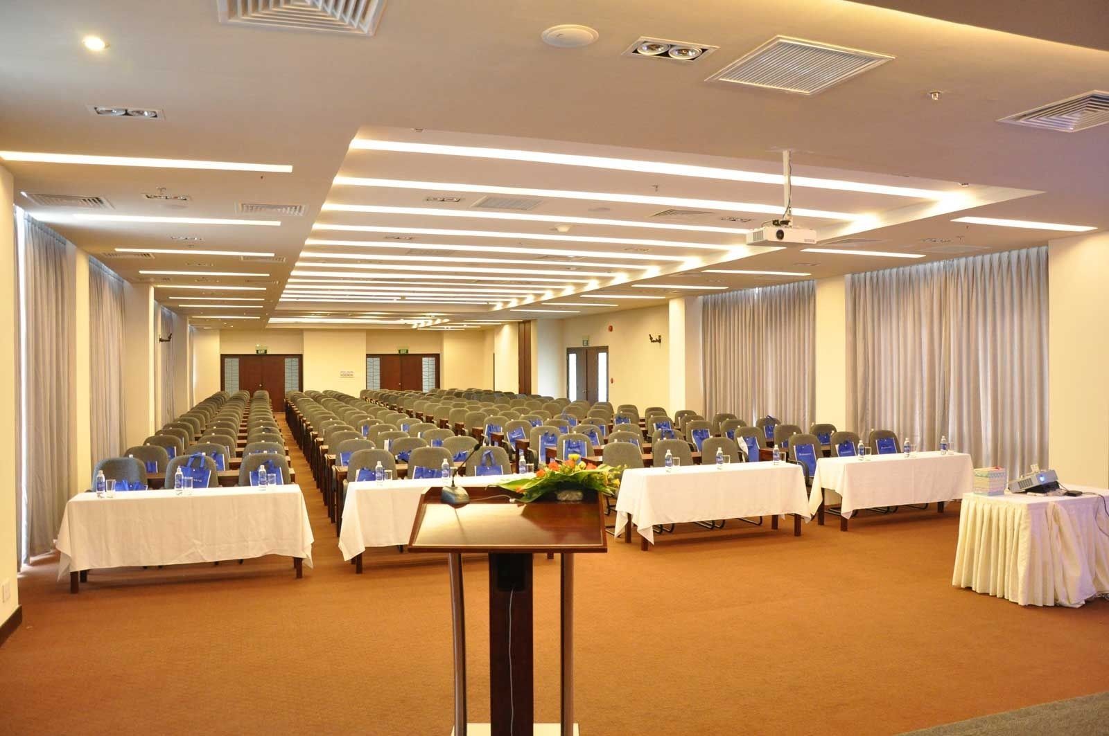 Un salón de conferencias con filas de sillas, mesas con botellas de agua y un proyector sobre una mesa.
