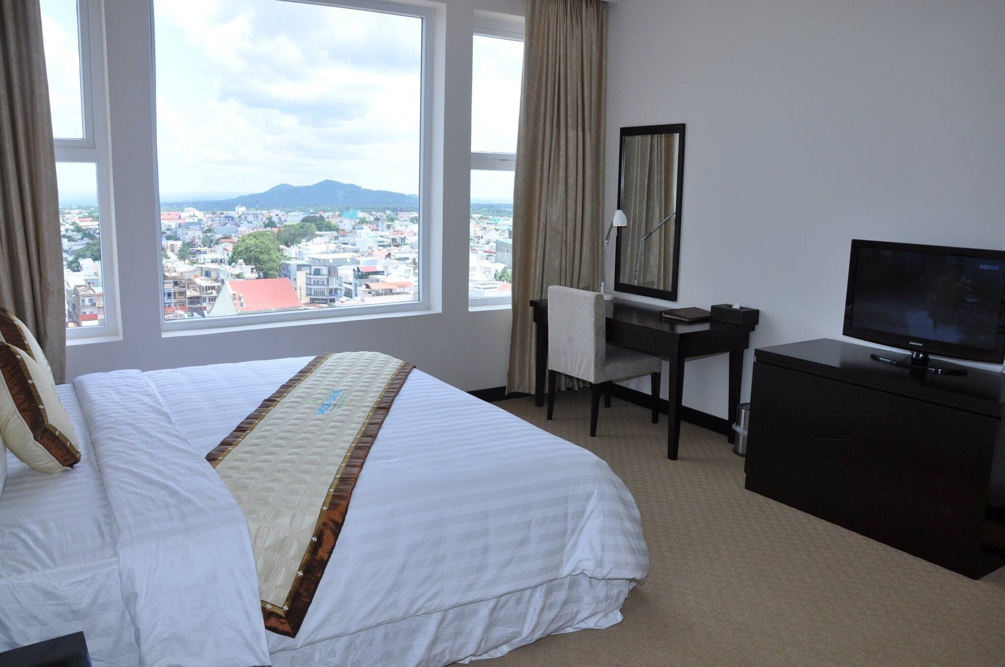 Una habitación de hotel con una cama grande, un escritorio, un televisor y vistas de la ciudad a través de amplios ventanales.