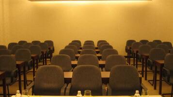 Uma sala de conferências com fileiras de cadeiras e mesas, equipada com garrafas de água e copos sobre as mesas.
