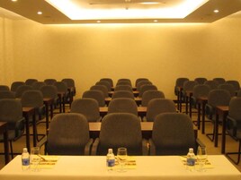 Uma sala de conferências com fileiras de cadeiras e mesas, equipada com garrafas de água e copos sobre as mesas.