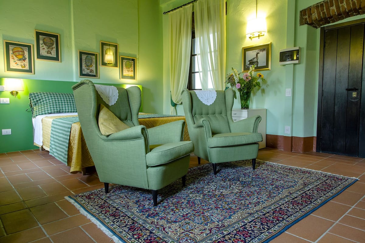 A room with two green armchairs, a bed with a checkered blanket, a patterned rug, and framed pictures on the wall.