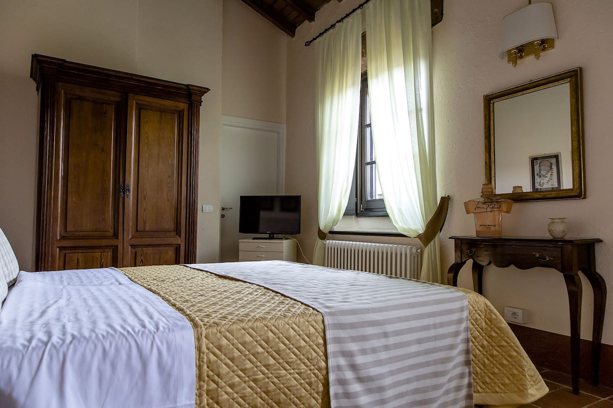 A bedroom with a bed, a television, a window with curtains, a radiator, and a mirror on the wall.