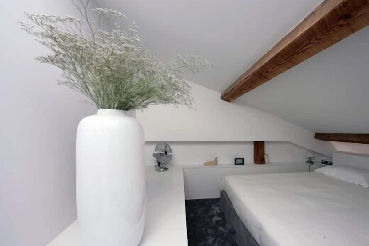 Un dormitorio moderno con un jarrón blanco grande lleno de flores, una cama con ropa de cama blanca y una viga de techo de madera.