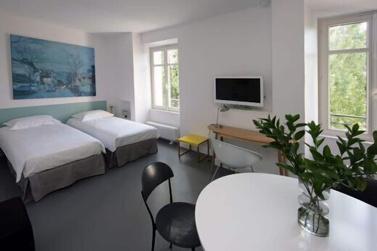 Habitación de hotel con dos camas, un escritorio con computadora, una silla, un televisor y una ventana con vista a follaje.