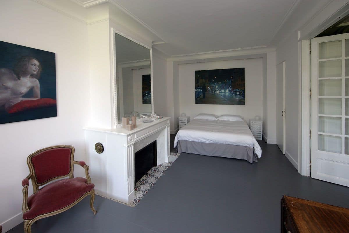 Un dormitorio con chimenea blanca, una cama, un sillón rojo y dos cuadros en las paredes.