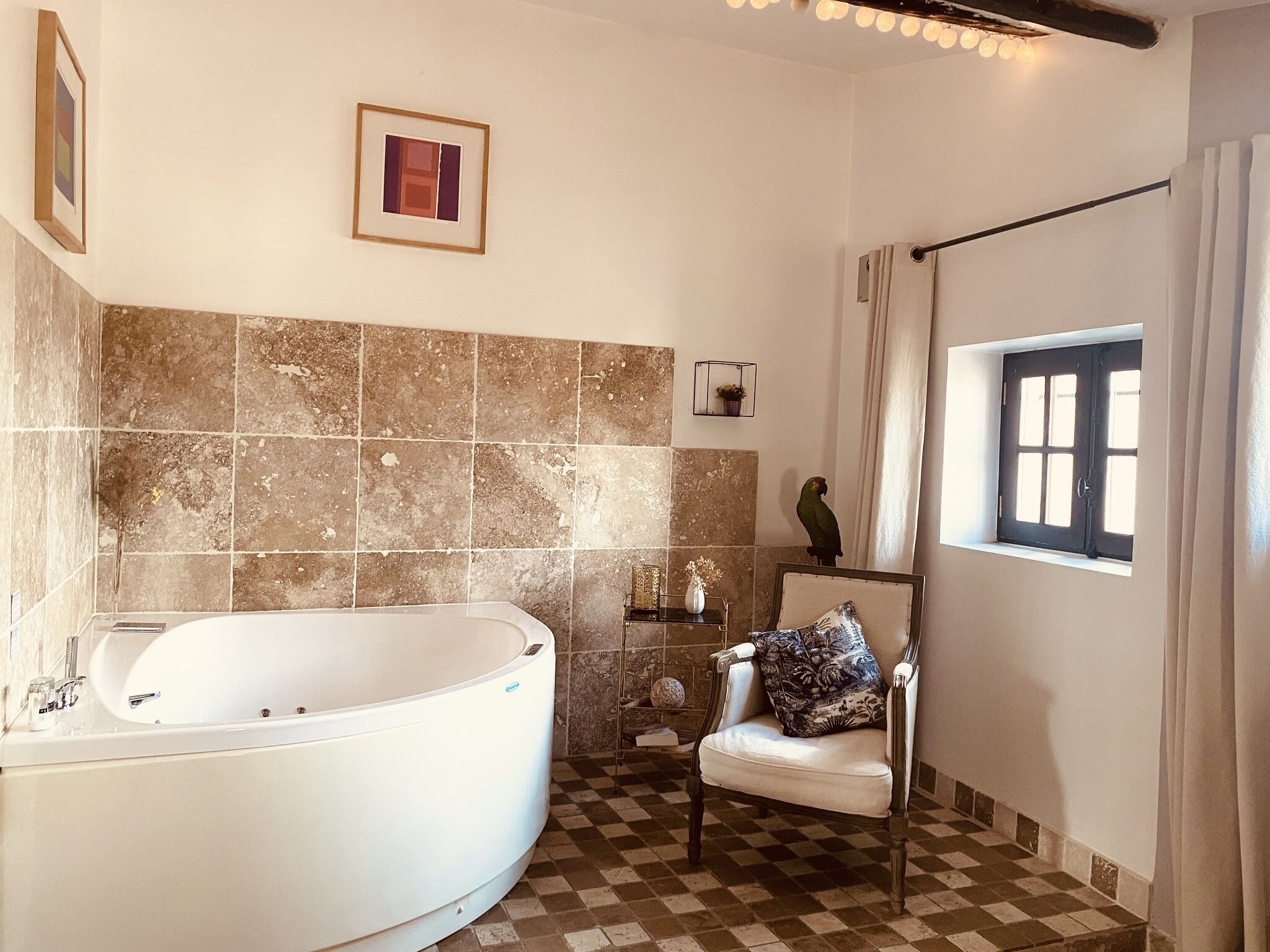 Un baño con una bañera independiente, pisos de baldosas de cuadros, un ventanal con cortinas y un cuadro en la pared.