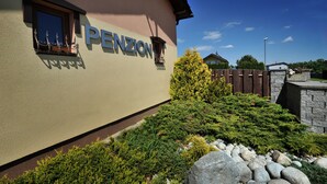A building with the name "PENZION" on its facade, surrounded by landscaped greenery and a stone wall.