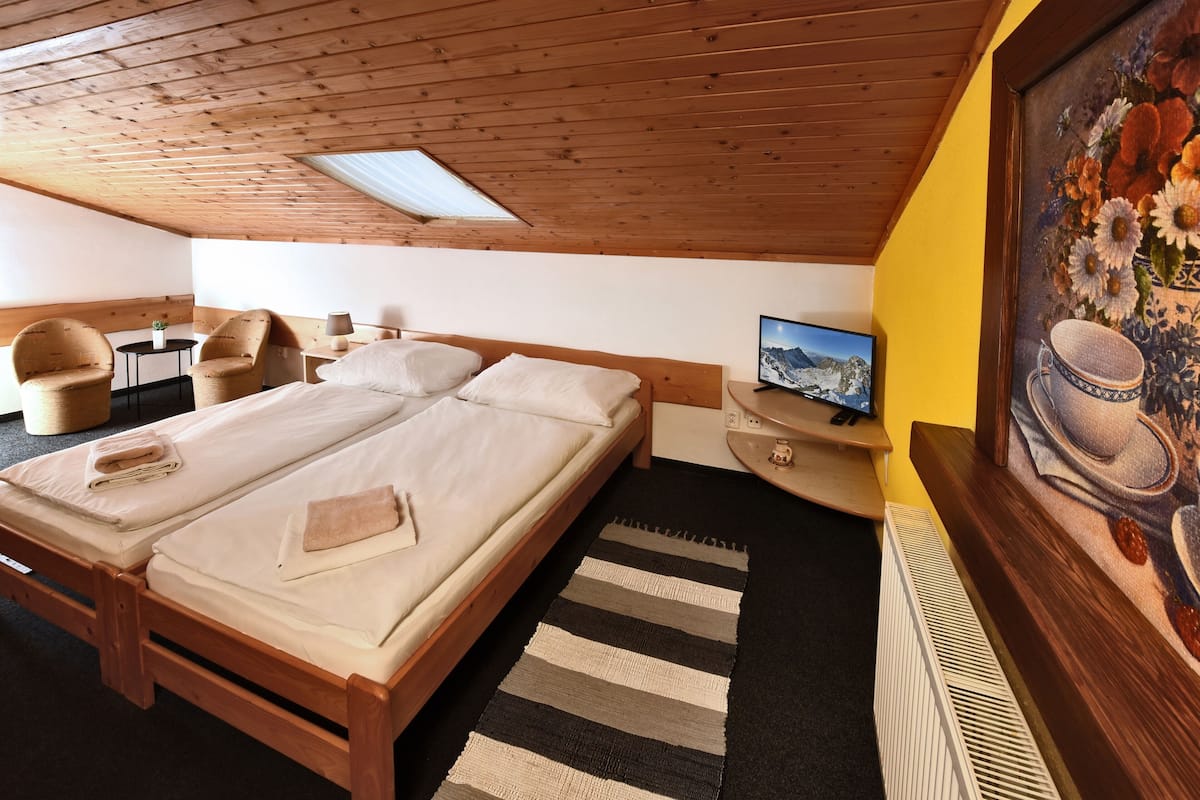 Una habitación con techo de madera, una cama con ropa de cama color beige, un televisor sobre un mueble de madera, un cuadro en la pared y una mesita con una planta.