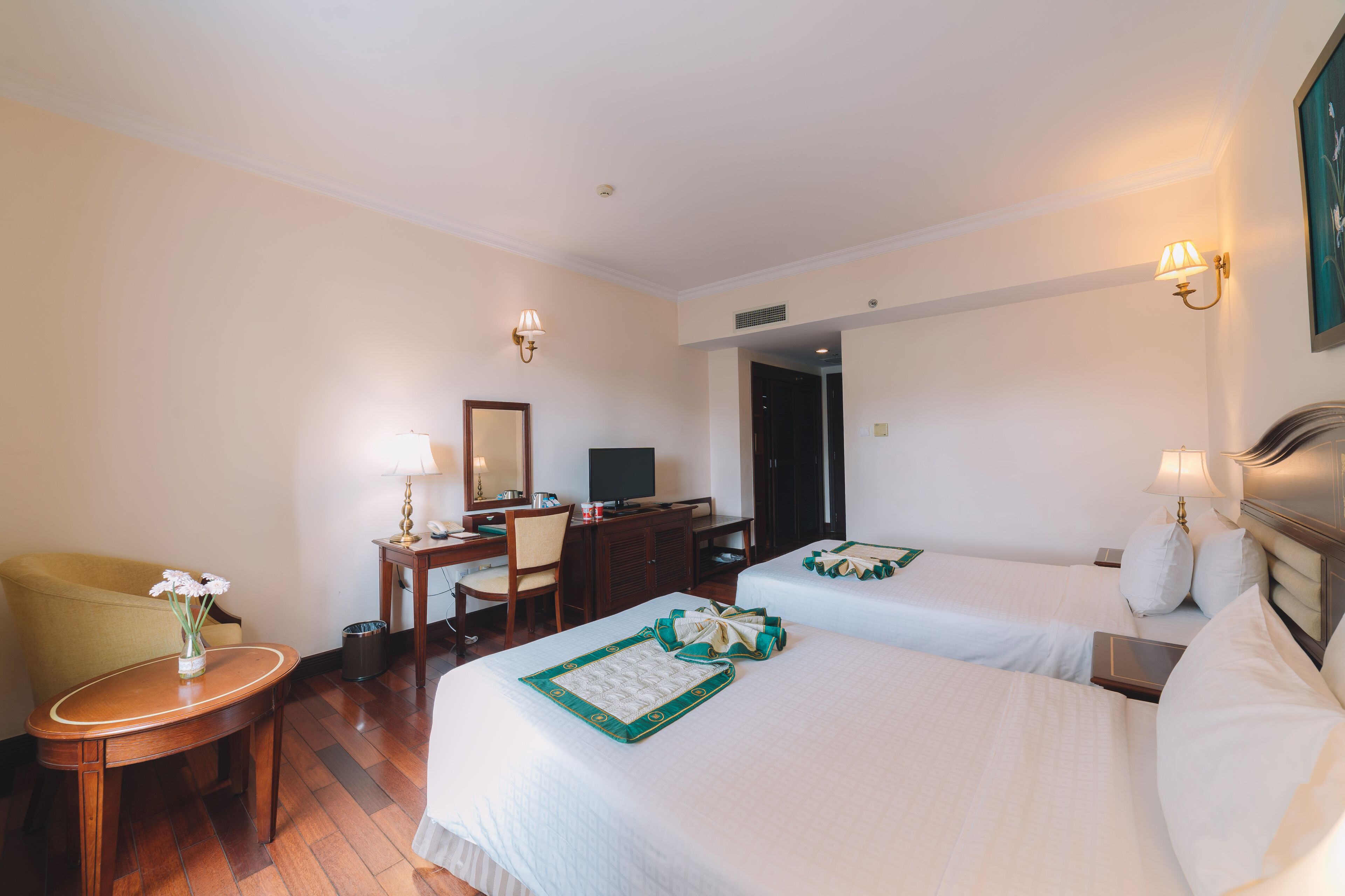 Habitación de hotel con dos camas, un escritorio, una silla, una mesita con un florero y un cuadro en la pared.