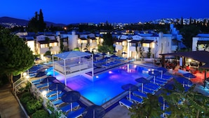 Piscina com espreguiçadeiras e guarda-sóis, cercada por edifícios iluminados.