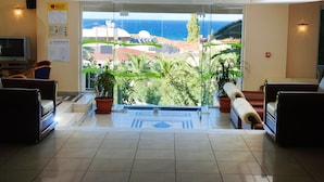 Un vestíbulo de hotel con puerta de cristal que da a un balcón con vista al mar y edificios. Hay plantas en macetas y un televisor a la izquierda.