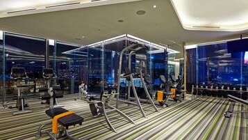 Une salle de sport moderne équipée de divers appareils d’exercice, de grandes fenêtres offrant une vue sur la ville et d’un sol à rayures.