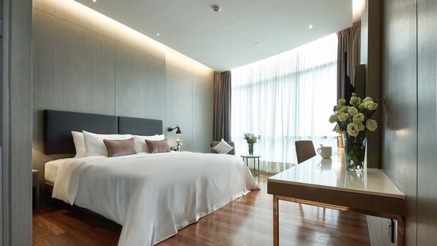 Una habitación de hotel moderna con una cama grande, mesitas de noche, una pequeña mesa con un jarrón de flores y un amplio ventanal con cortinas.