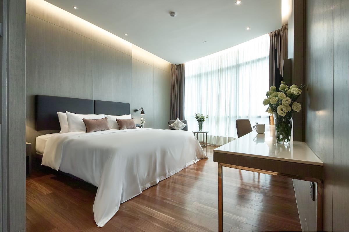 Una habitación de hotel moderna con una cama grande, mesitas de noche, una pequeña mesa con un jarrón de flores y un amplio ventanal con cortinas.