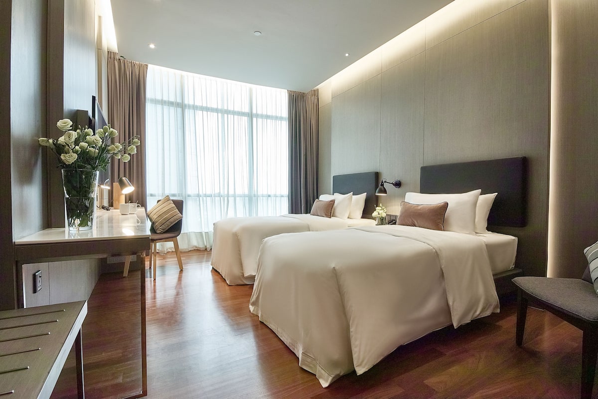 Una habitación de hotel con dos camas, un escritorio con silla, una lámpara y un florero con flores.