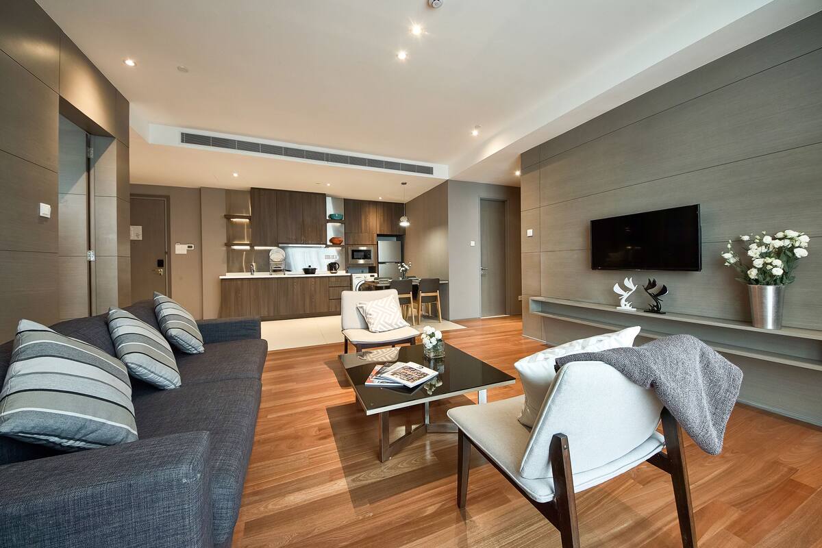 Una sala moderna con un sofá, una mesa de centro y un televisor. Se observa una cocina al fondo con una zona de comedor.