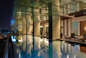 A modern hotel pool area with glass walls, lounge chairs, and a city view at night. - V E Hotel & Residence (Kuala Lumpur)