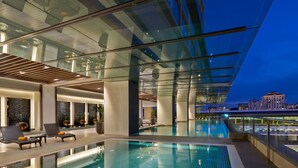 Una piscina interior moderna con paredes de vidrio, sillones de descanso y vistas a la ciudad al anochecer.