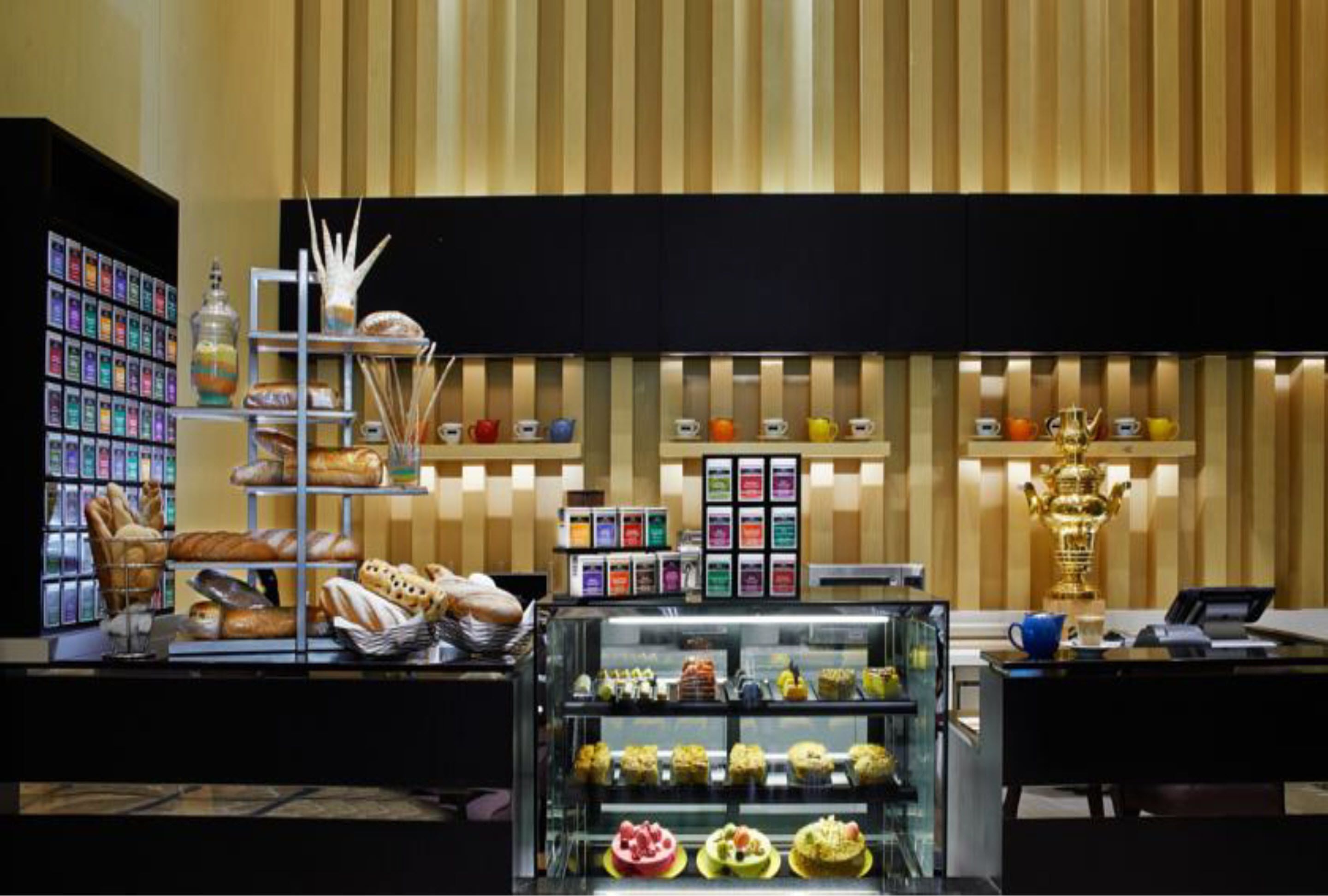 Vitrine de pastelaria com diversos doces e pão, estação de café com várias chávenas e uma máquina de café, e uma vitrine de vidro com sobremesas variadas.