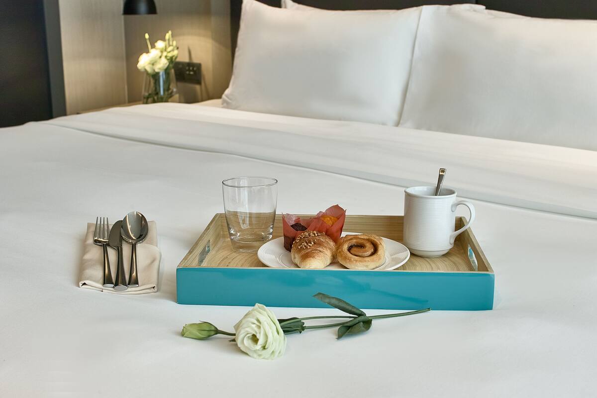 Una bandeja de desayuno ordenada sobre una cama, con una almohada blanca y un jarrón de flores al fondo.