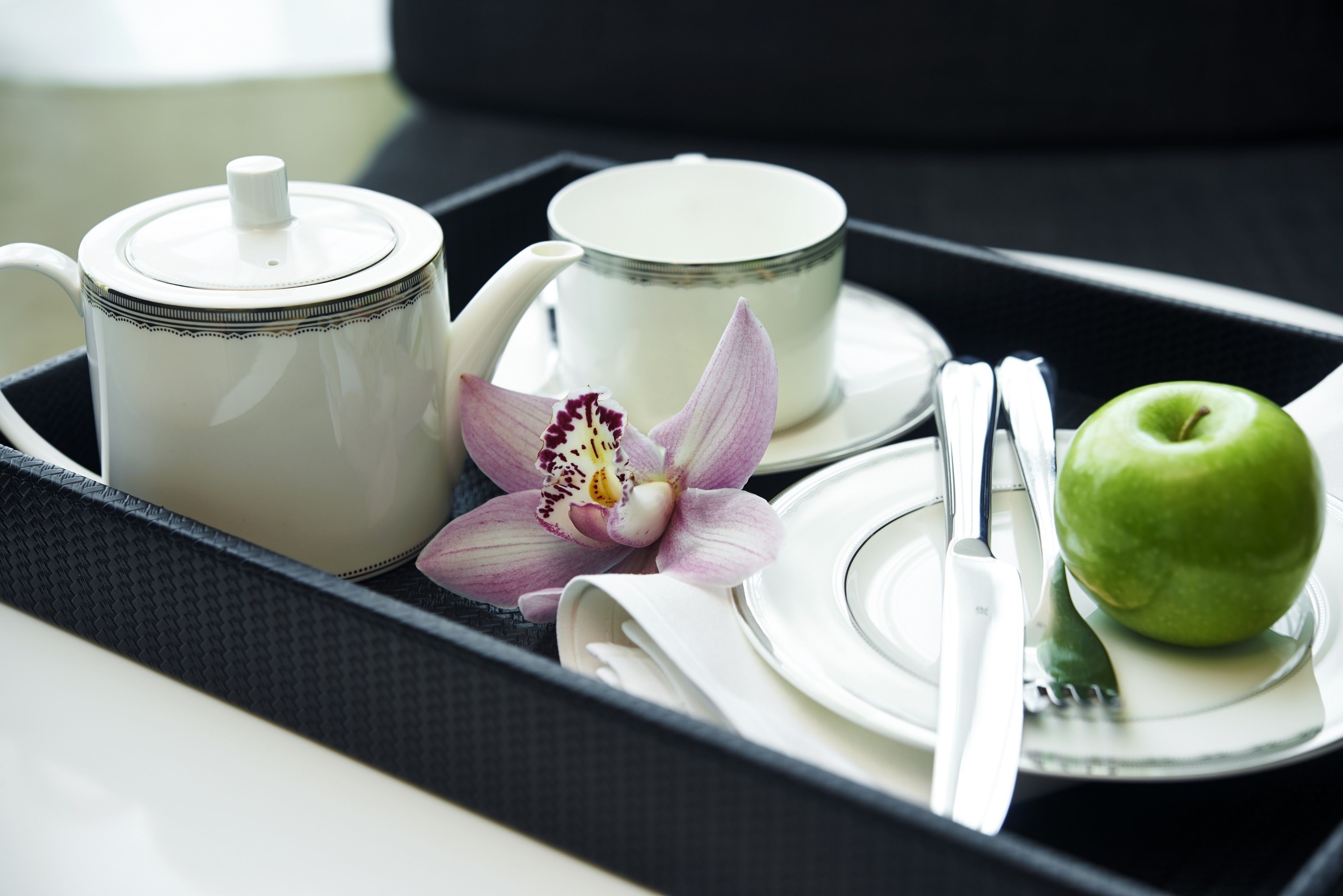 Una bandeja con tetera, taza, orquídea, manzana y cubiertos.