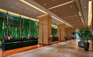 A modern hotel lobby with a reception desk, large windows with a green view, and a polished wooden floor.