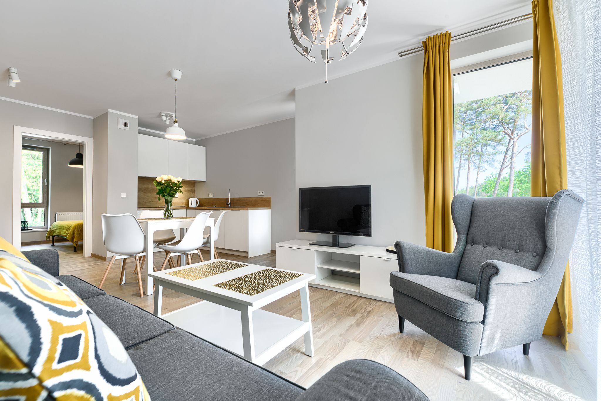 A modern living room with a grey sofa, a white coffee table, a grey armchair, a flat-screen TV, and a dining area with a white table and chairs.