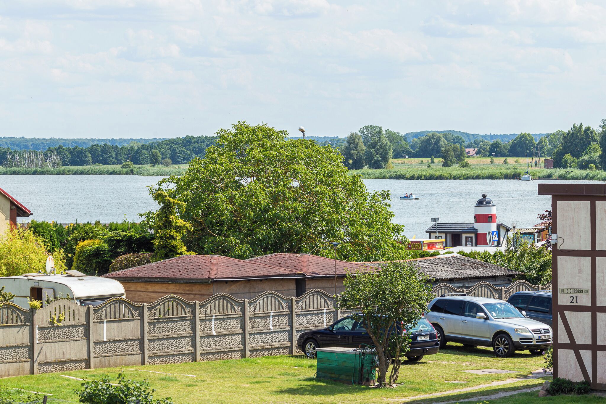 Een vakantiehuis aan het meer met een omheind erf, geparkeerde auto's en een vuurtoren op de achtergrond.