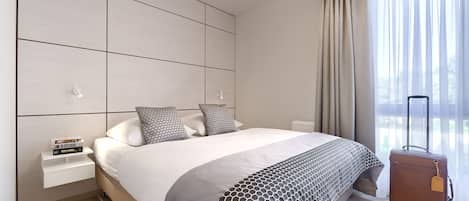 A modern hotel room with a large bed, a nightstand, a suitcase, and a view of the outdoors through sheer curtains.