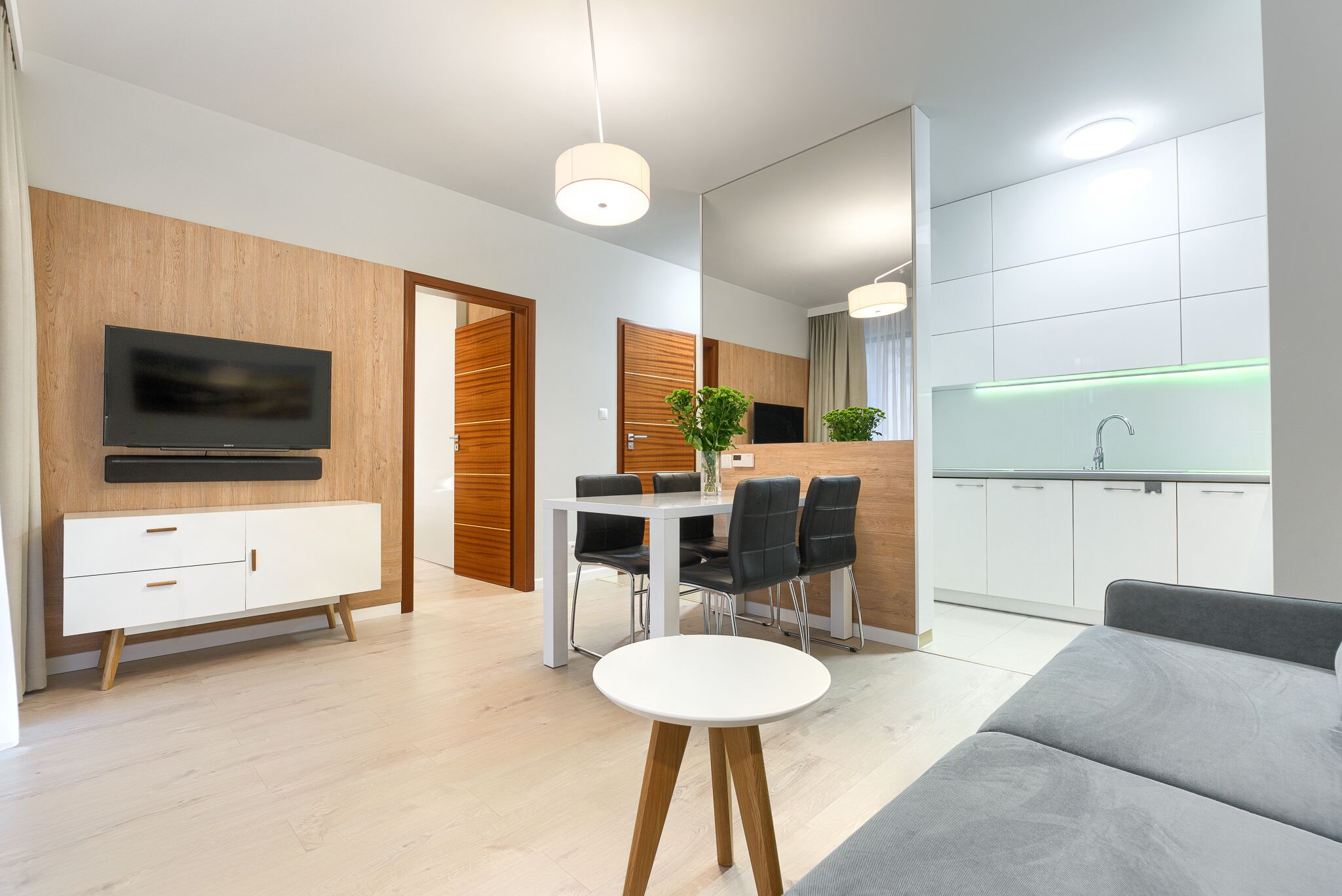 A modern living room with a flat-screen TV, a dining area with a white table and black chairs, and a kitchen with white cabinets and a green-lit backsplash.