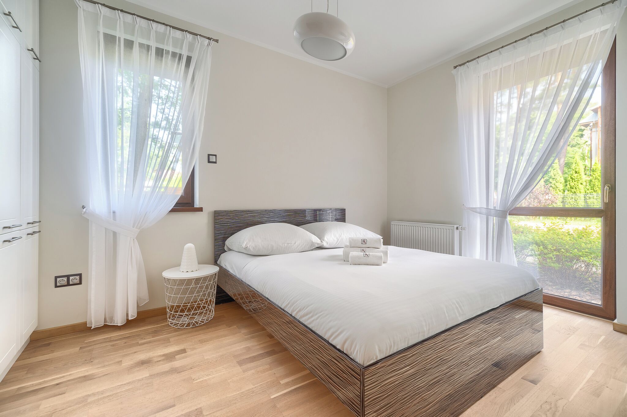 Un dormitorio moderno con estructura de cama de madera, ropa de cama blanca y cortinas traslúcidas.