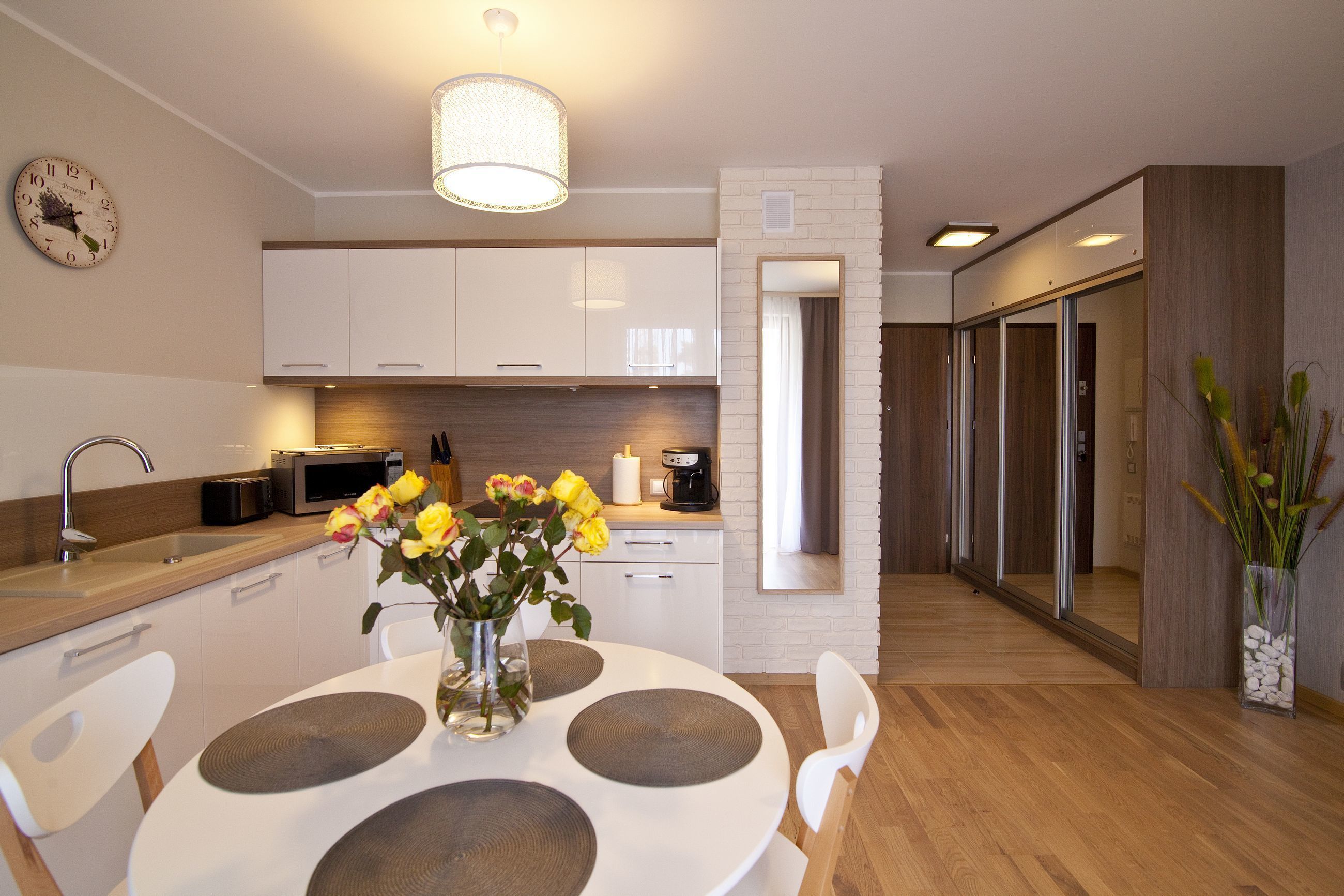 A modern kitchen with white cabinets, a microwave, and a sink. There's a dining table with a vase of yellow flowers.