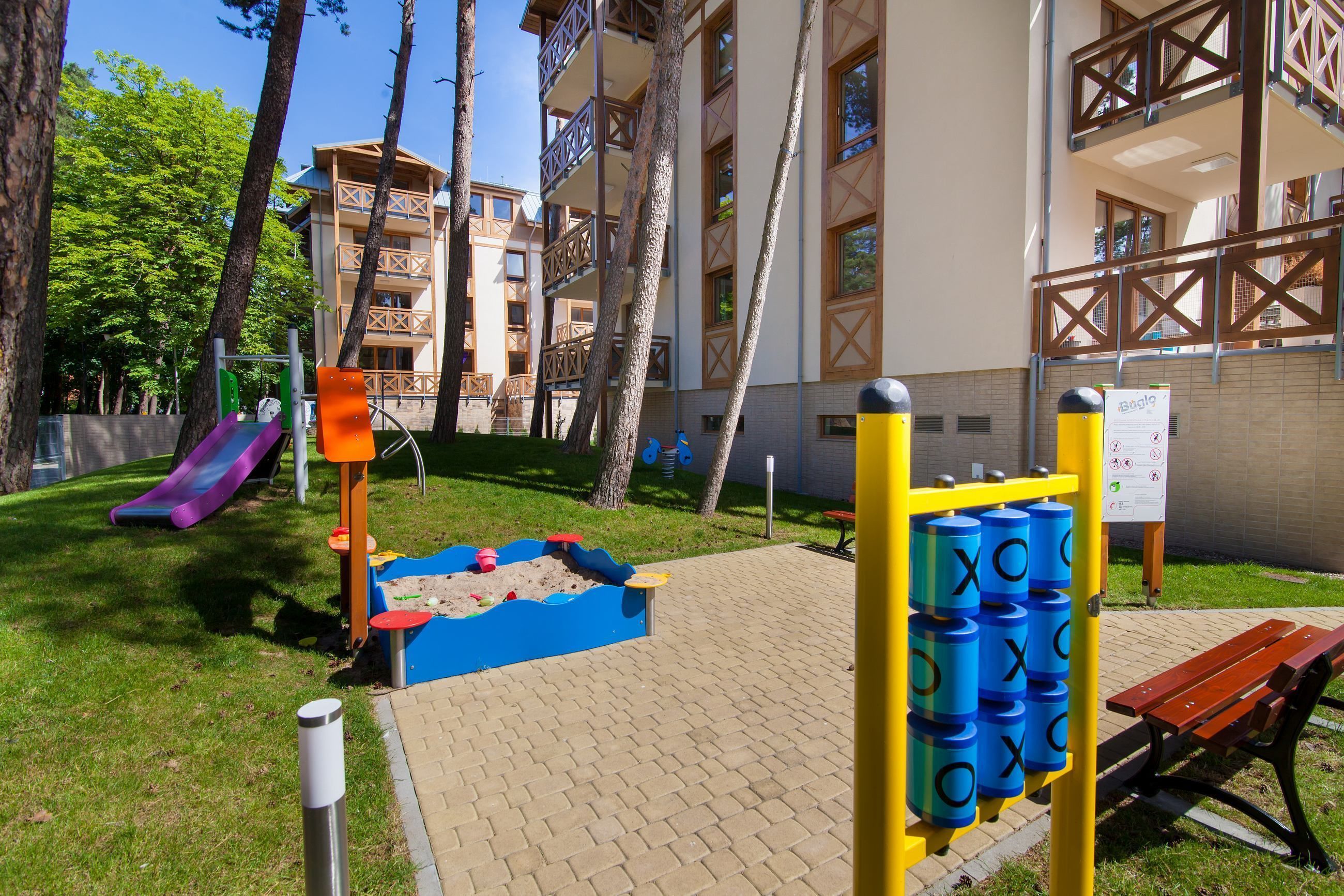 A playground with a slide, sandpit, and climbing structures, surrounded by trees and a building with balconies.