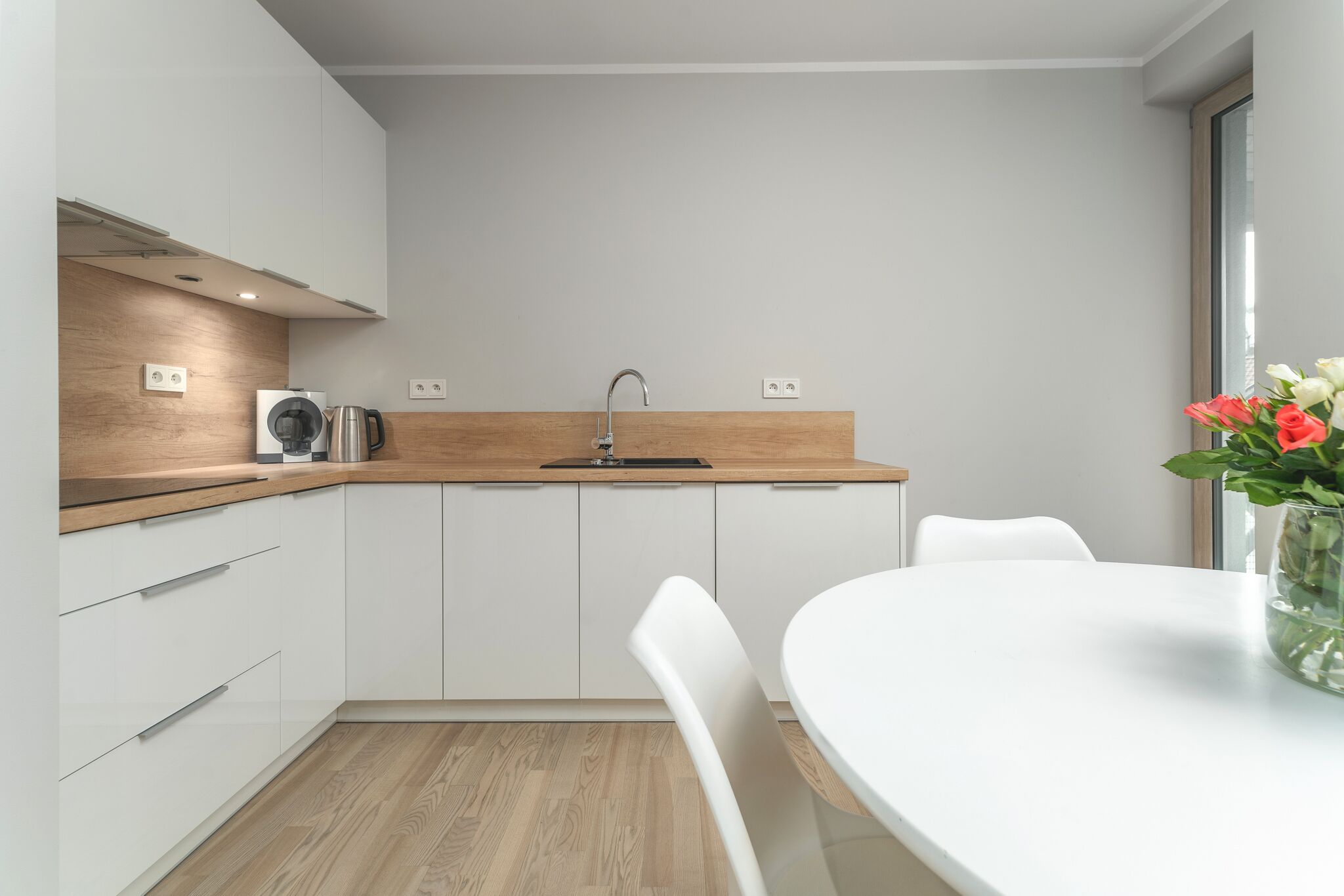 Una cocina moderna con armarios blancos, encimera de madera y un lavabo. Hay una mesa de comedor blanca con sillas y un florero con flores.