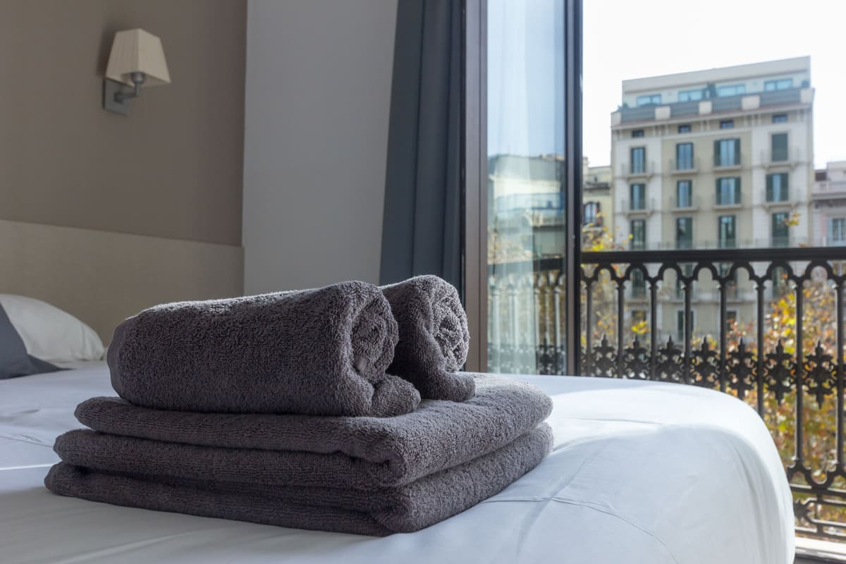 Een net opgemaakt bed met opgevouwen handdoeken en uitzicht op een stadsgezicht door een raam met een zwart ijzeren balkon.