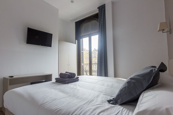 A hotel room with a bed, a TV mounted on the wall, a window with curtains, and a radiator.